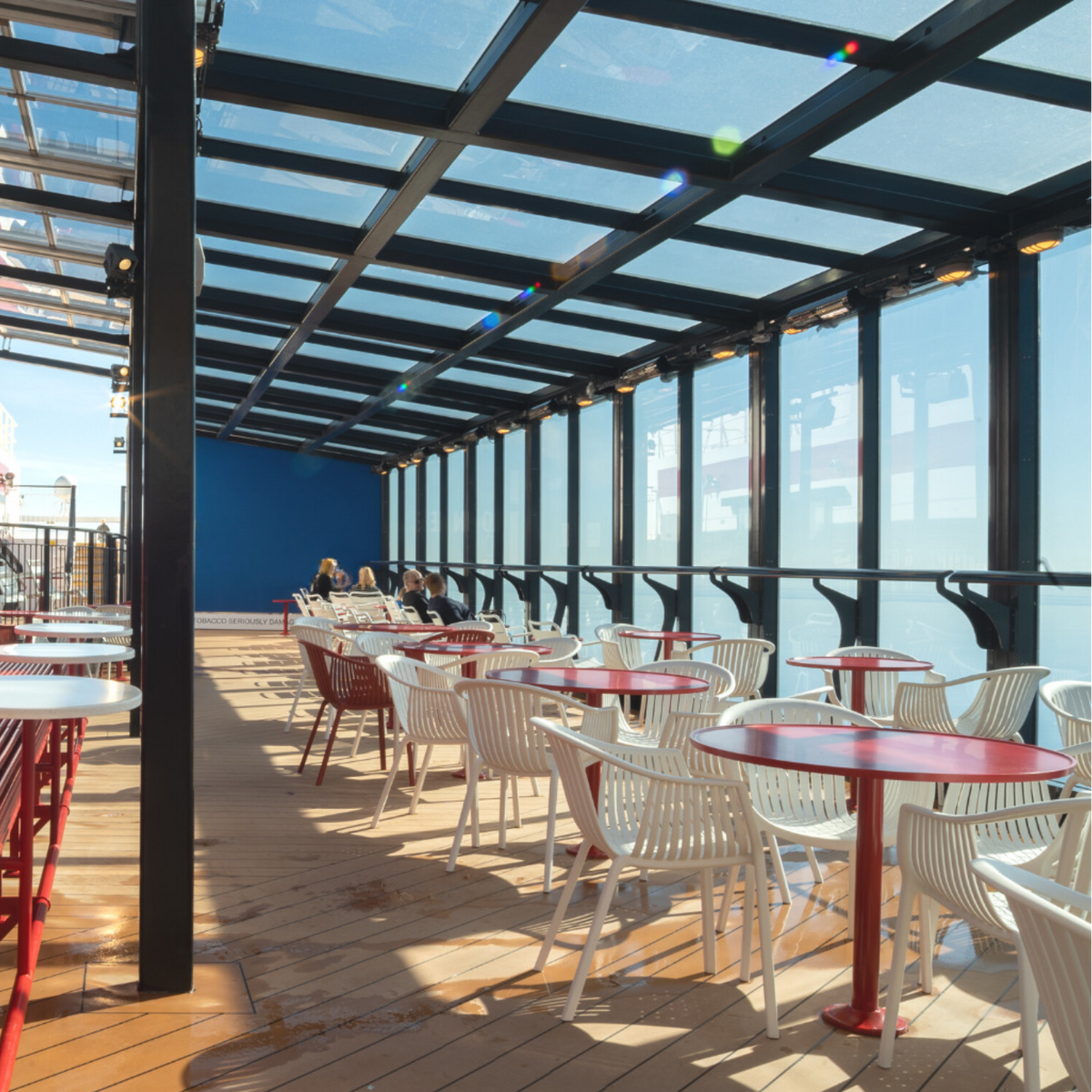 An outdoor dining area on a cruise ship with red round tables and white chairs arranged along a wooden deck