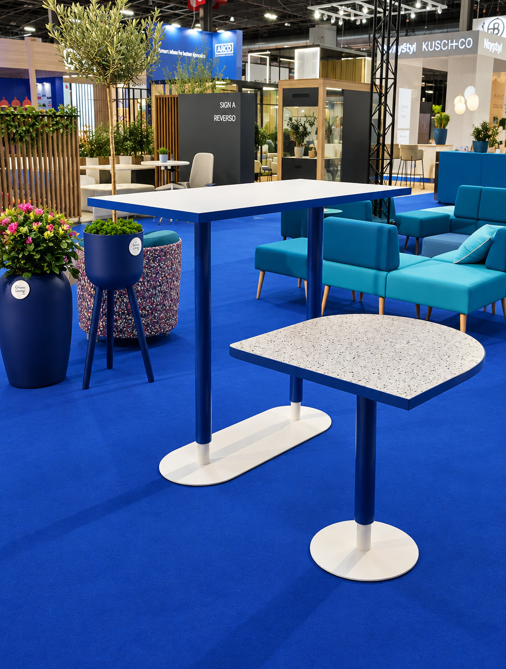 A modern exhibition hall setting featuring a tall rectangular table and a smaller oval table with blue frames and white circular bases, surrounded by turquoise chairs and a blue carpeted floor.